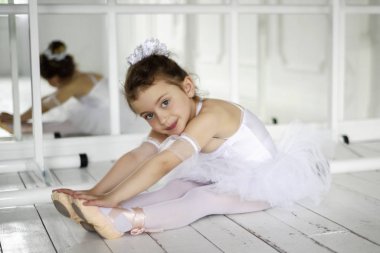 Klasik balerin çocuk prova odasında esneme hareketleri yapıyor. Arka planda aynaları olan 6 yaşında beyaz bale elbiseli bir kız yerde oturuyor. Çalışma oturumu