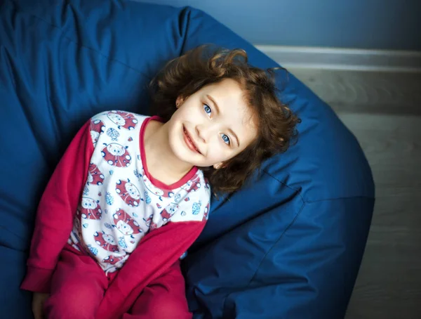 Niña de 6 años en pijama rosa acostada en otomana azul y mira hacia arriba con sonrisa. día de sueño. Felicidad, ocio y estilo de vida en la infancia. Luz natural del día — Foto de Stock
