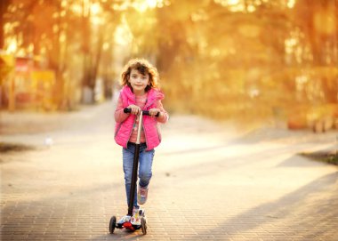 Küçük şirin 6 yaşında kot pantolon ve pembe yelek giyen bir kız scooter kullanıyor.