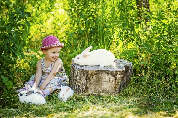 Niña de 3 años en vestido gris y sombrero rosa se sienta en el suelo con cuatro conejos alrededor. Un gran conejo de Pascua blanco se sienta en el tocón. El niño y el conejito se miran. Primavera Pascua en el jardín — Foto de Stock