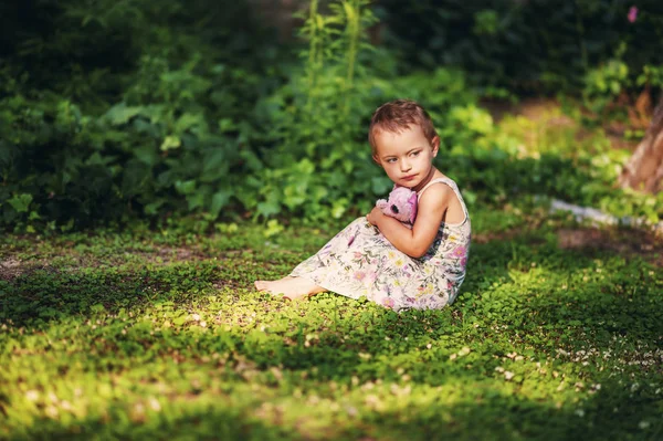 Маленькая 3-х летняя коротковолосая девочка в сером платье сидит на зеленой траве в саду с маленькой розовой игрушкой слона. Грустное дитя в летнее время. Аллергия. Закат. Естественное освещение — стоковое фото