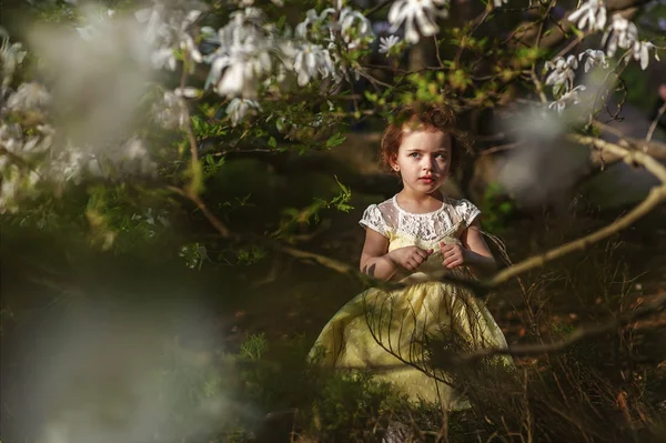 Piękna 5-letnia dziewczyna w żółtej sukience w kwitnącym ogrodzie magnolii. Wiosna - sezon alergii. Czas na zachód słońca. Kwiaty na gałęziach. Styl życia w ogrodzie botanicznym — Zdjęcie stockowe
