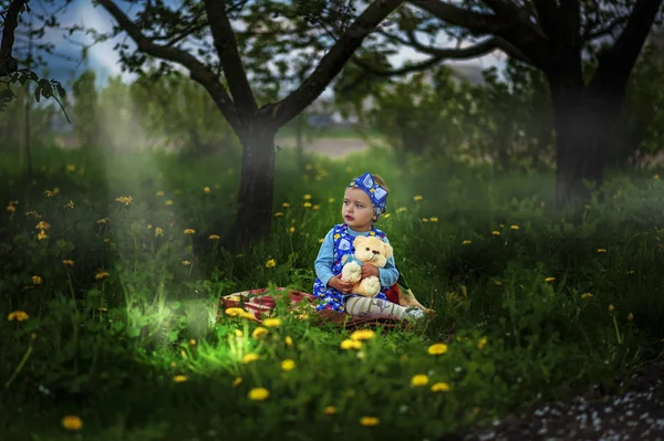 Mała 2-letnia dziewczynka siedzi na łące w ogrodzie w promieniach światła z kremowym niedźwiedziem zabawki w rękach. Wieczór. Wiosną. Potencjalnie uczulony. Dzieciństwo bliskie naturze. Żółte kwiaty dookoła — Zdjęcie stockowe