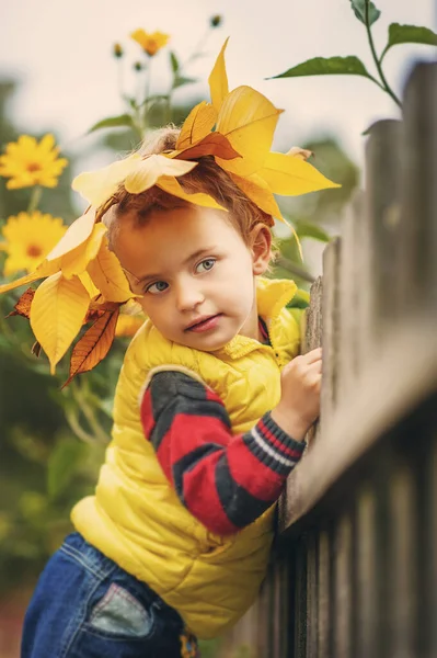 Das kleine Mädchen, 3 Jahre alt, mit einer gelben Weste und einem Kranz aus gelben Herbstblättern auf dem Kopf schaut weg. Kinderporträt im Herbst. Naturnah. Öko-Schönheit. Herbst. Kindheit. — Stockfoto