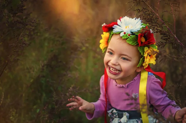 Όμορφο χαμογελαστό 3χρονο κορίτσι με ροζ φόρεμα και παραδοσιακό ουκρανικό στεφάνι στο κεφάλι στο ηλιοβασίλεμα. Χαμογελάστε στο πρόσωπο μικρό κορίτσι από την Ουκρανία. παραδοσιακά ρούχα και εορταστικές εκδηλώσεις — Φωτογραφία Αρχείου