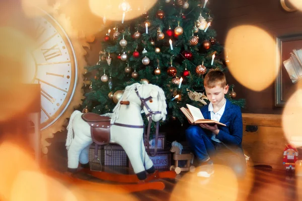 6 ans garçon assis sur le sol et lire le livre. Cheval à bascule et grande horloge à proximité. Veilles de Noël avec beau sapin de Noël et cadeaux. Enfant attendre des cadeaux dans la nouvelle année. Il y a des lumières. Casse-noisette — Photo