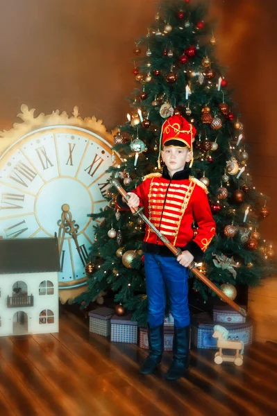 6 years old boy wearing like hussar with saber stay near christmas tree. Big clock and dollhouse on floor. Child in masquerade carnival costume of nutcracker. Dreams come true in christmas time