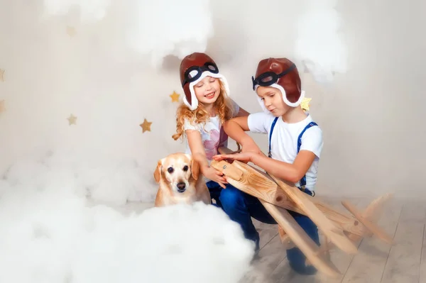 Brother and sister in aviator helmet playing with big wooden airplane in white clouds. Children and their dog together in sky with plane. Boy repair airplane. Girl help him. Happy childhood