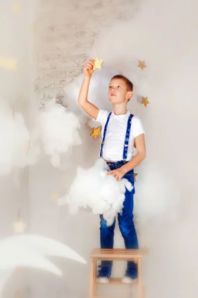 6 yaşında kot pantolon ve pantolon askısı giymiş bir çocuk bulutlarda merdivende durup yıldıza dokunuyor. Rüyalar çocuklukta gerçekleşir. Çocuklar için sihir. Çocuk dokunuşu yıldızı. Rüyada uç. — Stok fotoğraf