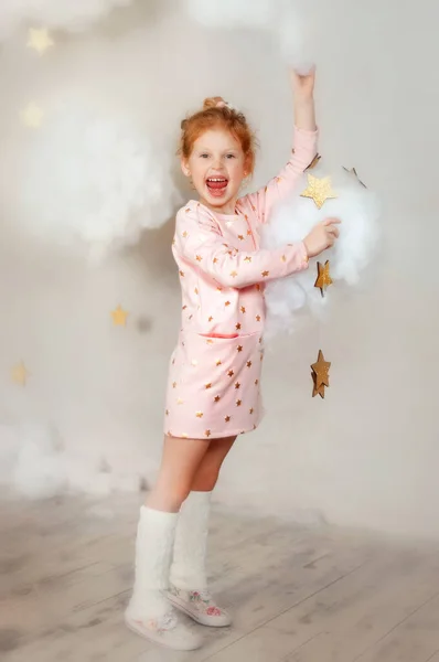 Chica de 5 años en vestido rosa con estampado de estrella y calcetines blancos se quedan en las nubes y las estrellas con una amplia sonrisa. Hermosa niña tocar las estrellas y el cielo. Sonriente sueño de niño soñador con magia — Foto de Stock