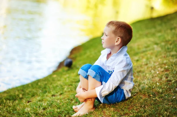 Ξυπόλητος 5χρονος κάθεται στη λίμνη και κοιτάει αλλού. Παιδί με λευκό πουκάμισο και μπλε τζιν χωρίς παπούτσια κοντά στο νερό, σκεφτείτε. Οργανικός οικολογικός τρόπος ζωής στην παιδική ηλικία — Φωτογραφία Αρχείου