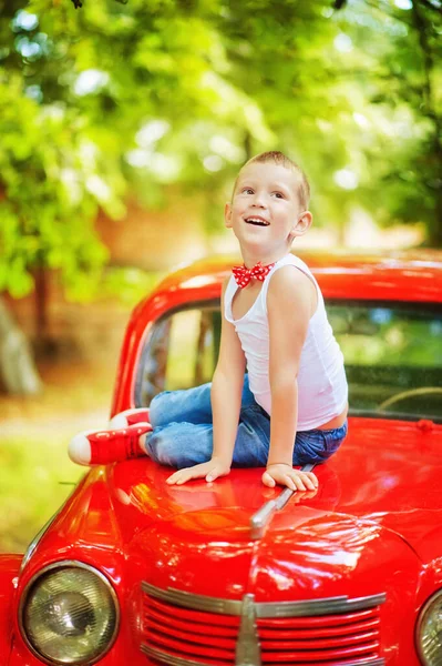 5 years old boy in blue jeans, white t-shirt and red bow tie sit on red retromobile old car and look up. Happy child outdoor. Vintage style. 60-s lifestyle. Stylish boy in summer clothes — Stock Photo, Image