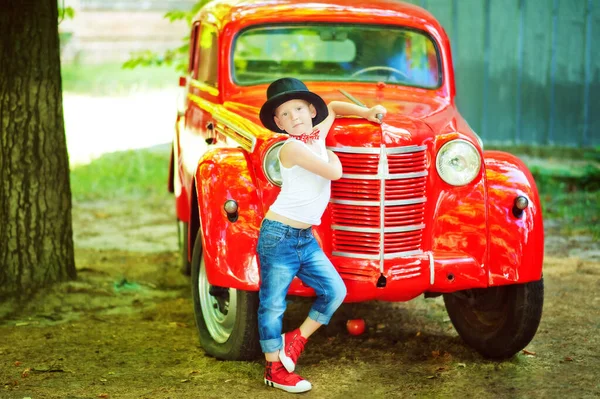 Pětiletý chlapec v modrých džínách, bílém tričku, červeném motýlku a černém válci se drží poblíž červeného retromobilního starého auta a dívá se do kamery. Šťastné dítě venku. Klasika ročníku. Životní styl 60s — Stock fotografie