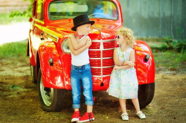 5-jähriger Junge in blauer Jeans, weißem T-Shirt, roter Fliege und schwarzem Zylinder und 3-jähriges Mädchen in heller Kleidung stehen neben rotem Retromobil-Oldtimer und schauen einander an. Glückliche Kinder im Freien — Stockfoto