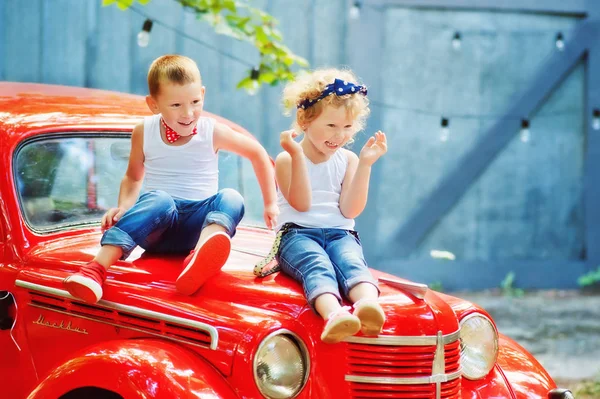 5 ετών αγόρι με μπλε τζιν, λευκό t-shirt, κόκκινο παπιγιόν και 3 ετών κορίτσι με μπλε τζιν και λευκό t-shirt κάθονται σε κόκκινο οπισθοκινητό παλιό αυτοκίνητο και χαμόγελο. Ευτυχισμένα παιδιά έξω. Παραδοσιακός τρόπος ζωής — Φωτογραφία Αρχείου