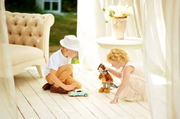 Zwei Kinder spielen auf dem Boden im Sommerpavillon im Garten. Bruder und Schwester verbringen Zeit miteinander. Junge mit weißer Sommermütze und Spielzeugauto. Mädchen im hellen Pfirsichkleid mit Mäusespielzeug. Geschwister spielen — Stockfoto