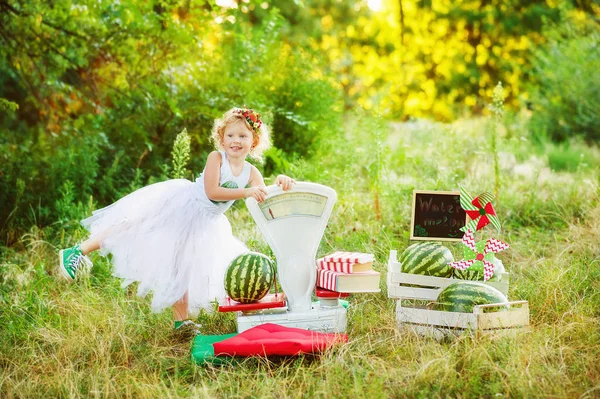 Pequeña niña de 3 años en falda de bailarina jugar en vendedor de sandía con grandes escalas blancas anticuadas. Niño emocional de pie cerca de grandes sandías en el parque. Alimento ecológico saludable para niños . — Foto de Stock