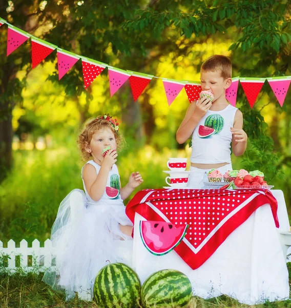 Dos niños hermano y hermana comen pastelitos dulces en un hermoso picnic decorado. Niños hambrientos niño y niña comiendo al aire libre. Qué hacer en verano con los niños. Camisetas decoradas con sandía — Foto de Stock