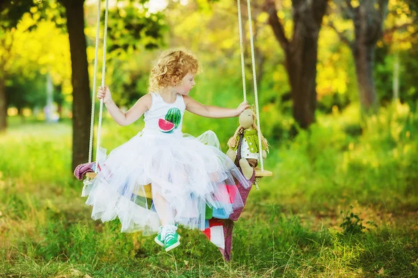 Dreijähriges Mädchen in grünen Turnschuhen, weißem Ballerinas-Rock und weißem T-Shirt mit Wassermelonenaufdruck schwingt mit ihrem Spielzeug im Garten. Schaukel im Freien für Kinder. Kind spielt im Sommer im Park. — Stockfoto