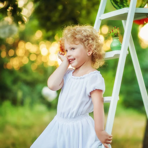 Πορτρέτο του 3 ετών μικρό σγουρό ξανθό όμορφο κορίτσι με το ένα χέρι κοντά στο πρόσωπό της. Παιδί στο φως ηλιοβασιλέματος μείνετε στη σκάλα στον κήπο. Χώρος για κείμενο. Μικρό κορίτσι έκπληξη στο πάρκο — Φωτογραφία Αρχείου
