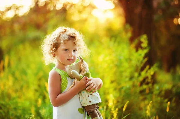 Портрет 3 лет маленькая кудрявая блондинка красивая серьезная девушка с игрушкой в руках. Ребенок при солнечном свете остается в саду. Пространство для текста. Маленькая игривая девочка в парке. ребенок рядом с природой . — стоковое фото