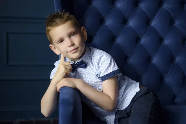 7 yaşında, mavi papyonlu, sinsi gülüşlü ve yüzünün yanında elleri olan beyaz bir çocuğun portresi. Modaya uygun küçük çocuk mavi koltukta oturur ve kameraya bakar. Metin için boşluk — Stok fotoğraf