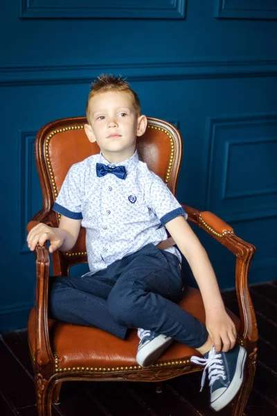 Mavi papyonlu, mavi spor ayakkabılı, kahverengi deri sandalyede oturan 7 yaşındaki beyaz bir çocuğun portresi. Şık görünümlü küçük çocuk sandalyeye otur ve kameraya bak. — Stok fotoğraf