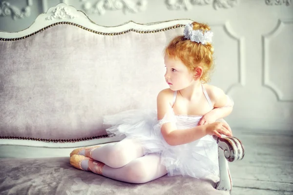 Retrato pequeña bailarina rizada rubia de 4 años sentada en la sala de luz en el sofá en el vestido de ballet tutú blanco y pointes. Relajación en el estudio de ballet. Retrato en estilo vintage. Bailar es un trabajo duro — Foto de Stock