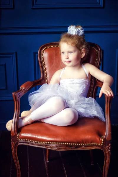 Retrato pequeña bailarina rizada rubia de 4 años sentada en habitación oscura en silla de cuero marrón en vestido de ballet tutú blanco y pointes. Relajación en el estudio de ballet. Bailar es un trabajo duro — Foto de Stock