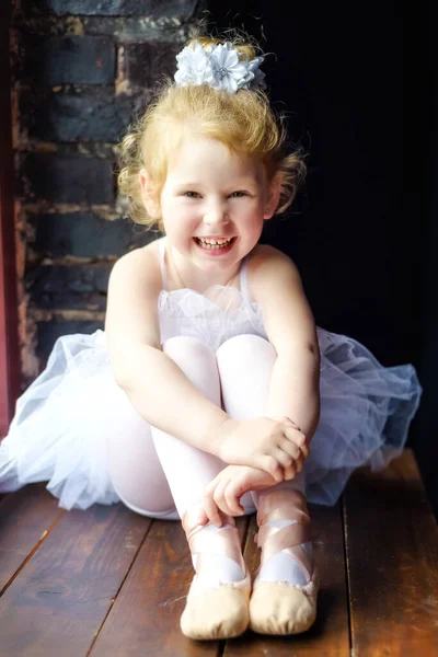 Retrato pequeña 4 años rubia rizada bailarina sonriente sentada en la ventana en habitación oscura en vestido de ballet tutú blanco y pointes. Relajación en el estudio de ballet. Pausa en el entrenamiento. Bailar es un trabajo duro — Foto de Stock