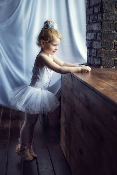 Retrato pequeña bailarina rizada rubia de 4 años de edad de pie cerca de la ventana en habitación oscura en vestido de ballet tutú blanco y pointes. Relajación en el estudio de ballet. Pausa en el entrenamiento. Bailar es un trabajo duro —  Fotos de Stock