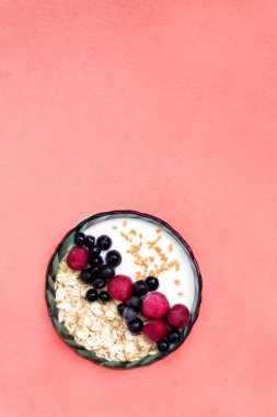 Sağlıklı organik vejetaryen kahvaltısında dikey manzara, bir kase müsli ve yoğurt taze çilek, frenk üzümü ve kirazlı canlı mercan rengi arka planda sol köşede.
