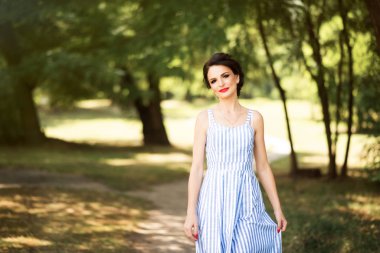 Yazın parkta kırmızı ruj sürmüş mavi ve beyaz çizgili kolsuz elbiseli güzel esmer kızın portresi. Yaz modası yaşam tarzı. Doğaya yakın rahatla..