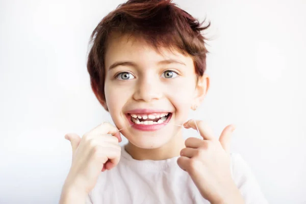 7 yaşındaki beyaz tişörtlü gülümseyen çocuğun yatay portresi beyaz arka planda dişlerini diş ipiyle temizliyor. Çocukluktan kalma sağlık ve diş bakımı. Diş ipinin nasıl kullanılacağını. Metin için boşluk. — Stok fotoğraf