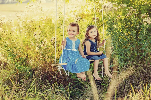 2 meninas em vestido azul no balanço no verão no campo. Crianças felizes brincando ao ar livre ao pôr do sol no verão. Crianças descalças alegres e ativas perto da natureza. Ácaros harving perigosos na grama — Fotografia de Stock