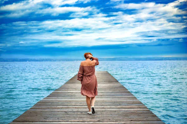 Back of faceless red haired woman in red dress walking by wooden pier on Garda lake in cloudy day. Italian vacation. Garda lake great tourist destination and attraction in North Italy.