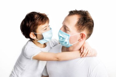 Child and dad in blue medical masks and white t-shirts hugging and looking at each other on white background. Coronavirus protection. Medical care. Illness covid-2019 prevention