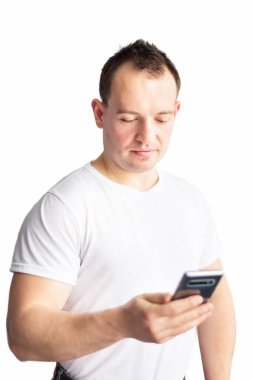Beyaz tişörtlü yakışıklı, kendine güvenen beyaz bir adamın dikey portresi beyaz arka planda cep telefonundan mesaj okuyor. Evden iletişim ve iş. Modern dünyada iş.