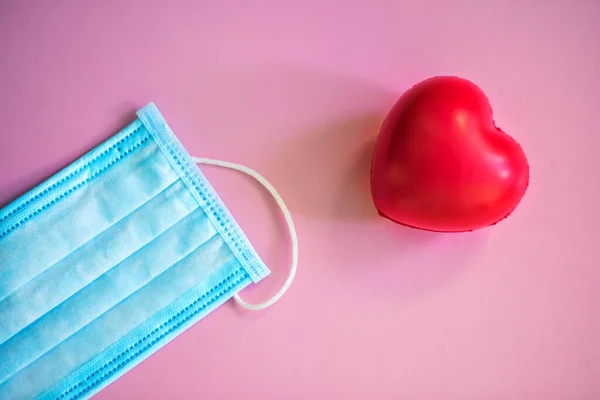 Medical face mask and red heart. Medical protective mask on pink background with red heart nearby. Disposable surgical face mask. Healthcare and medical concept. Protection from coronavirus covid-2019