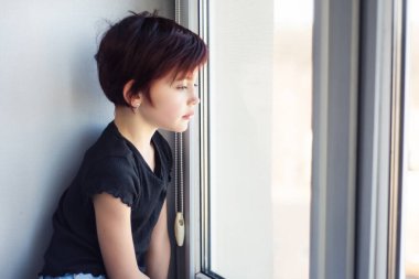 Pencereden bakan üzgün küçük kız. Çocuk karantina sırasında, pencereden dışarı bakan koronavirüs salgınından dolayı. İzolasyon ve yalnızlık.