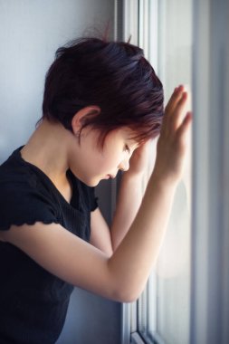 Pencereden dışarı bakan, cama elle dokunan üzgün küçük kızın dikey resmi. Çocuk karantina sırasında pencereden aşağıya bakan koronavirüs salgınından dolayı. İzolasyon ve yalnızlık.