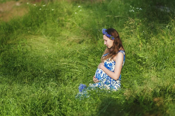 夏天的日落时分 孕妇头戴花环 穿着蓝色衣服 坐在青草丛中的垂直肖像 怀孕和接近大自然的健康的有机生活方式 — 图库照片