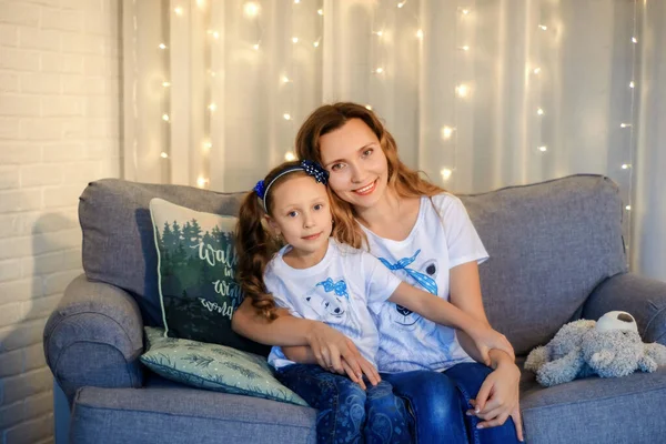 Sweet little girl together with beautiful young mom sitting on sofa in living room at home in evening. Happy family concept. Love. Trust. Togetherness. Parents and children. Family time. Stay home.