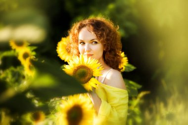 Ayçiçeği tarlasında arka planda sarı elbiseli güzel kızıl saçlı bir kadının yakın plan portresi. Yaz kırsal konsepti. Doğa tatiline yakın. Kadın ve ayçiçekleri. Yaz ışığı