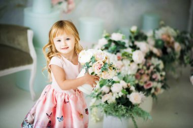 Parlak stüdyoda, pembe elbiseli, elinde çiçekli bir hediye kutusu olan, 3 yaşında, gülümseyen küçük bir kızın portresi. Evin içinde küçük bir çocuğun fotoşopu var. Anneler Günü.