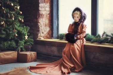 Kahverengi eski moda şapkalı ve elbisesi olan 4 yaşındaki güzel, kıvırcık, sarışın kız Noel ağacının yanındaki pencere pervazında el ısıtıcısı ile oturuyor. Çocuklu klasik kartpostal