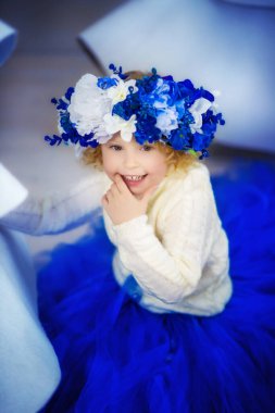 Küçük, gülen, 4 yaşında, kalın kazaklı, mavi etekli, mavi ve beyaz çiçekli büyük yapay çelenkli sarışın bir kızın dikey portresi. Baharı bekliyorum. Kış portresi. Gülümseyen çocuk