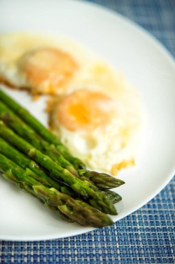 Fırında pişmiş taze kuşkonmaz resmi. Parmesan peynirinin altında kızarmış yumurta ve mavi arka planda büyük beyaz tabak, üst manzara. Mevsimlik bahar besin konsepti. Diyet olsun. Mevsimlik.
