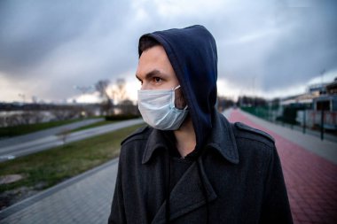 Genç adam dışarıda koruyucu tıbbi maske takıyor. Karantina sırasında dışarı çıkıyor. Koronavirüs yüzünden sosyal mesafeler. Pandemik koruma konsepti. Bulutlu bir arka plan. COVID-19