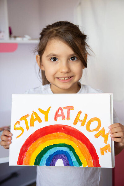 Stay at home due coronavirus pandemic concept. Little girl paints a rainbow on stay at home poster. Chase the rainbow flashmob. Positive activities during quarantine and staying at home. COVID - 2019.